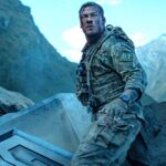 Alan ritchson s 81 standing next to part of the destroyed machine and looking beat up in war machine.jpeg
