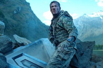Alan ritchson s 81 standing next to part of the destroyed machine and looking beat up in war machine.jpeg