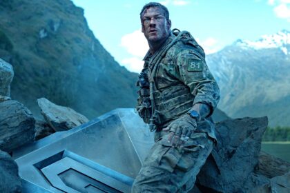 Alan ritchson s 81 standing next to part of the destroyed machine and looking beat up in war machine.jpeg